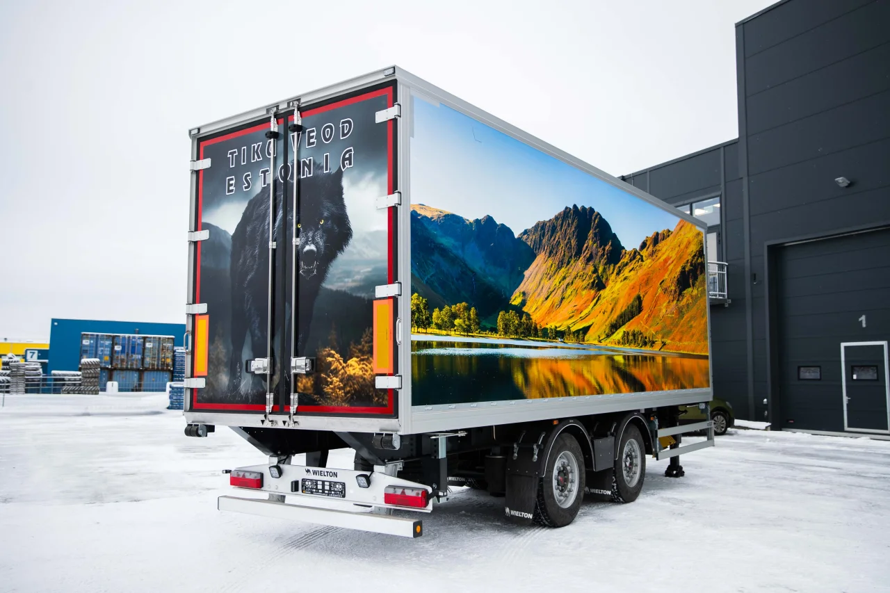 A side view of a large white semi-trailer parked on a snowy ground. The side of the trailer features a massive, high-definition wrap of a scenic mountain landscape with golden-brown slopes reflecting in a calm lake under a clear blue sky.