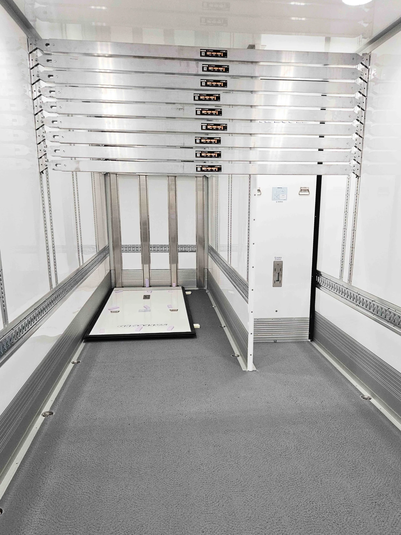 Interior view of a white cargo trailer showing a specialized load-securing system. Multiple adjustable horizontal aluminum bars are stacked against the front wall, with vertical tracking rails lining the sides and a grey anti-slip floor.
